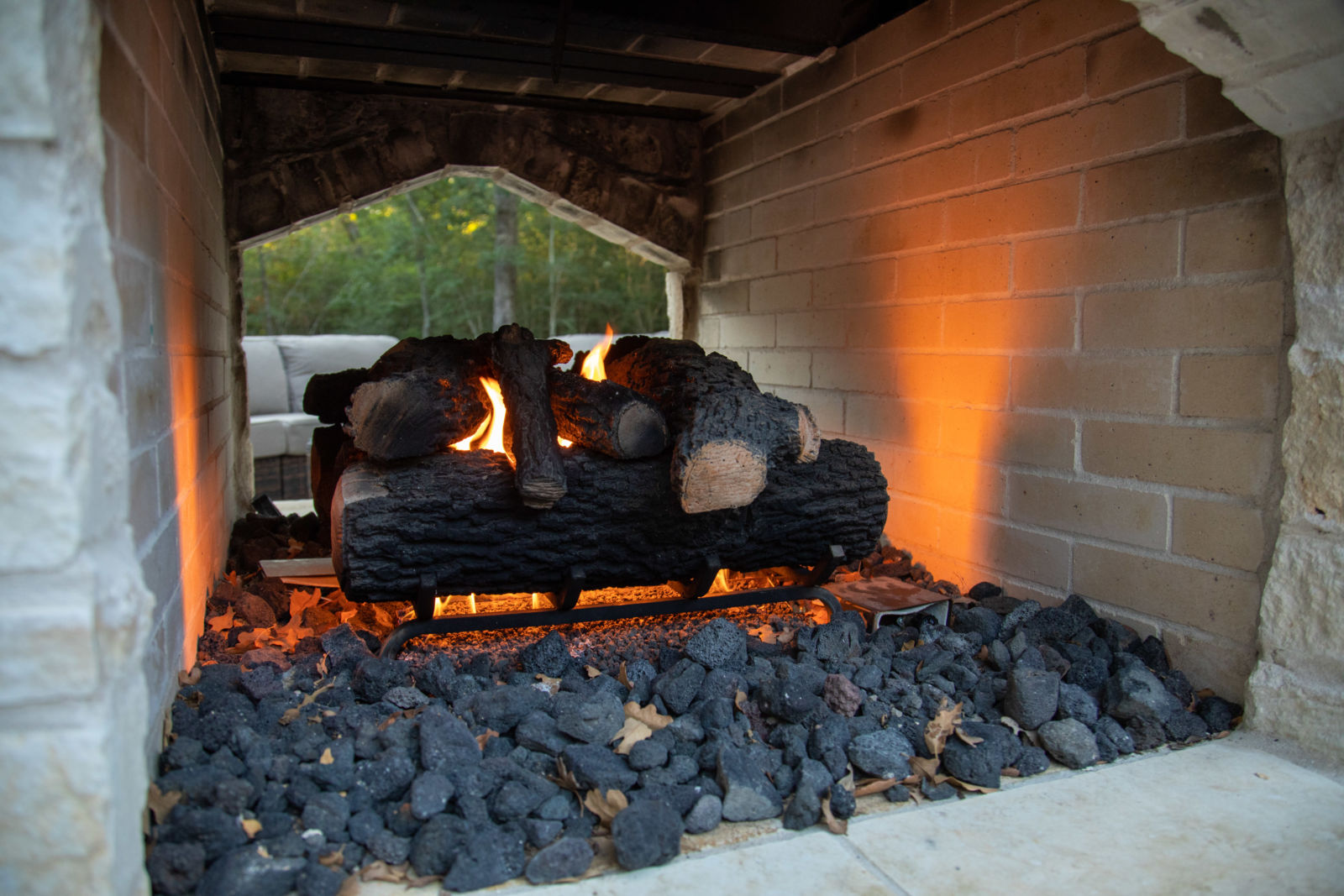 alt="logs in outdoor fireplace with glowing fire on black rocks, couches off in distance in background"