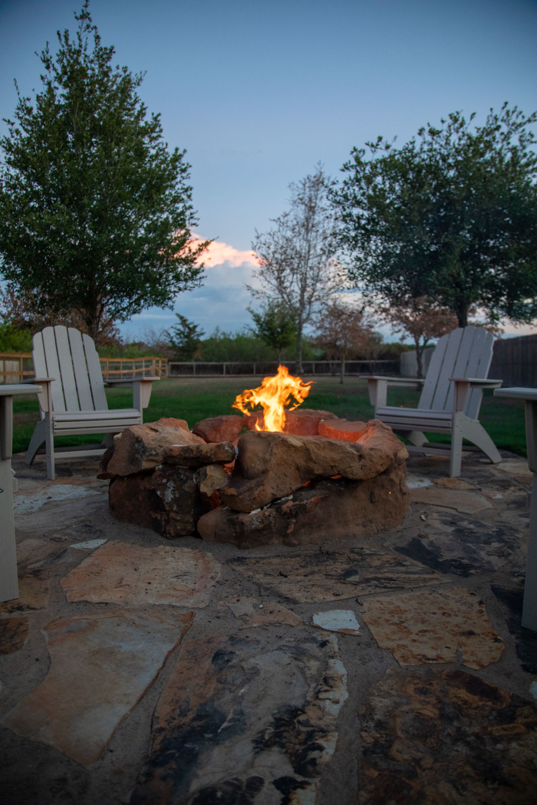 alt="outdoor fireplace with fire in fire pit in the evening surrounded by chairs"